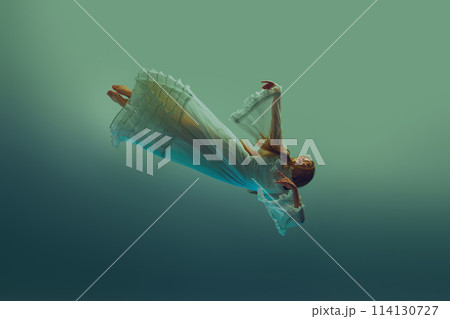 Woman in white vintage dress falls slowly through serene aqua background, evoking sense of weightless descent. 114130727