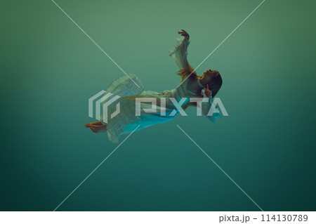Woman in ruffled dress appears suspended in time and space, she gracefully poised against serene aqua background. Woman in ruffled dress appears suspended in time and space, she gracefully poised against serene aqua background. 114130789