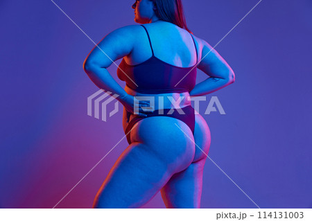 Cropped image of female body in underwear, young girl with plus size body posing against purple studio background in neon light Cropped image of female body in underwear, young girl with plus size body posing against purple studio background in neon light 114131003