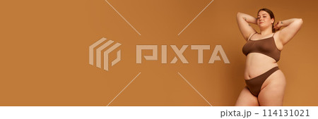 Young woman with oversized body in cotton underwear posing against light brown background. 114131021