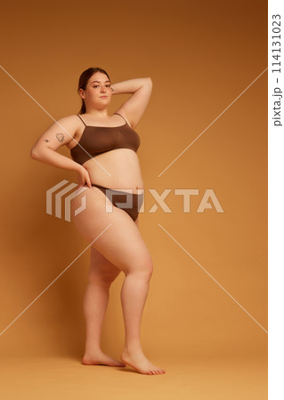 Full-length image of beautiful young girl feeling confident in her natural, plus size body, standing in underwear against light brown studio background 114131023