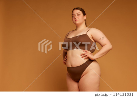 Confidence and grace. Beautiful young girl with plus size body posing in cotton underwear against light brown studio background 114131031