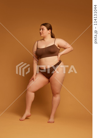 Full-length image of beautiful young girl feeling confident in her natural, plus size body, standing in underwear against light brown studio background 114131044