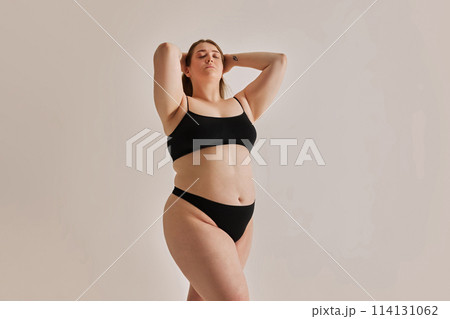 Serene expression of self-love. Young elegant girl with plus size body shape posing in underwear against grey studio background 114131062