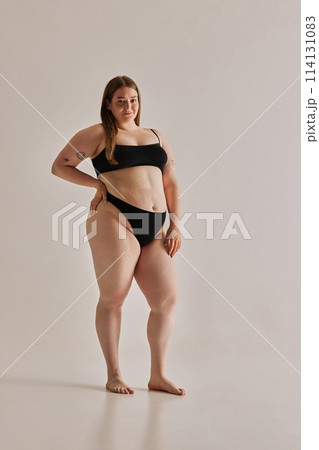 Full-length image of beautiful young girl with chubby body shape posing in black underwear against grey studio background Full-length image of beautiful young girl with chubby body shape posing in black underwear against grey studio background 114131083