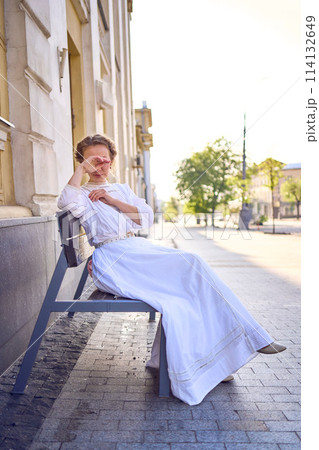 elegant middle age woman in a white vintage dress sitting on a bench in the morning city elegant middle age woman in a white vintage dress sitting on a bench in the morning city 114132649