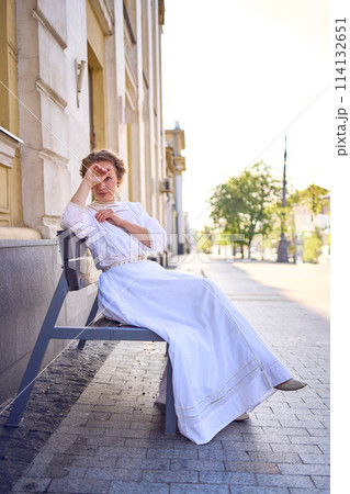 elegant middle age woman in a white vintage dress sitting on a bench in the morning city 114132651