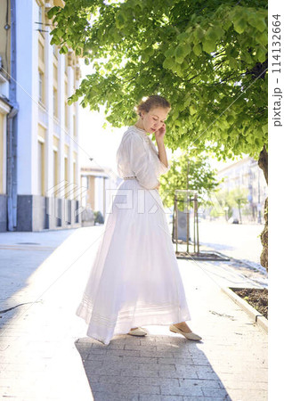 elegant middle age woman in a white vintage dress against the background of historical buildings in the morning light 114132664