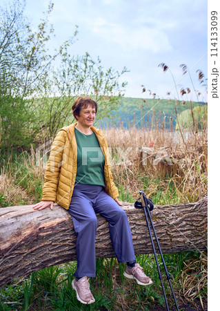 elderly woman rests on the river bank after Nordic walking exercise 114133099