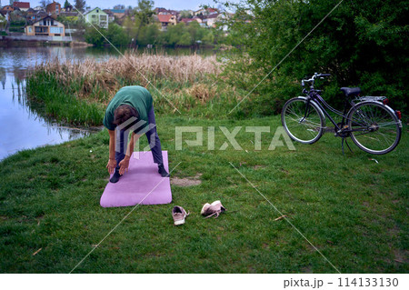 old woman doing exercises in the morning on the river bank, a bicycle and a yogamat next to her 114133130
