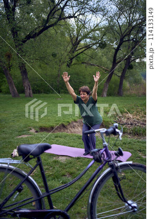 old woman practices yoga in the morning on the river bank, came from home by bicycle old woman practices yoga in the morning on the river bank, came from home by bicycle 114133149