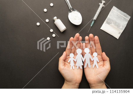 Protect family from drug addiction. Hands holding paper cut of family with drug powder, syringe and pills on dark background. International Day against Drug Abuse. Protect family from drug addiction. Hands holding paper cut of family with drug powder, syringe and pills on dark background. International Day against Drug Abuse. 114135904