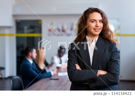 Confident caucasian businesswoman stands in office, arms crossed. Blonde boss oversees team meeting, embodies female leadership. Modern office with diverse staff discussing strategies. Confident caucasian businesswoman stands in office, arms crossed. Blonde boss oversees team meeting, embodies female leadership. Modern office with diverse staff discussing strategies. 114138422
