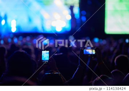 Fans record live music at evening outdoor fest with smartphones, stage lights illuminate excited audience, performers entertain on stage, energetic atmosphere youthful joy, festive celebration. Fans record live music at evening outdoor fest with smartphones, stage lights illuminate excited audience, performers entertain on stage, energetic atmosphere youthful joy, festive celebration. 114138424