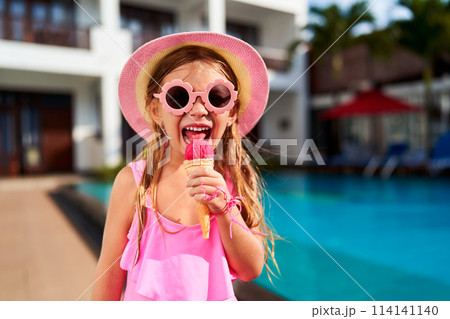 Cheerful girl in pink tank top and trendy sun hat enjoys red ice cream cone by poolside. Child cools off on sunny day, smiling. Summer kids fashion, leisure by water in warm weather. Happy moment. Cheerful girl in pink tank top and trendy sun hat enjoys red ice cream cone by poolside. Child cools off on sunny day, smiling. Summer kids fashion, leisure by water in warm weather. Happy moment. 114141140