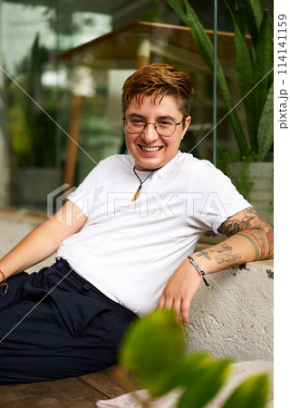 Smiling transgender person sits casually, modern coworking space with plants in background. Gen Z Individual with tattoos break exudes confidence, inclusion atmosphere. Casual smart creative office. Smiling transgender person sits casually, modern coworking space with plants in background. Gen Z Individual with tattoos break exudes confidence, inclusion atmosphere. Casual smart creative office. 114141159