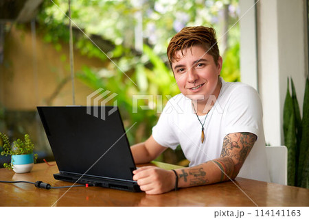 Confident transgender pro works on laptop in eco-friendly office. Smiling, tattooed, casual wear, tech-savvy. Workplace equality, LGBTQ pride, modern workspace with plants. Confident transgender pro works on laptop in eco-friendly office. Smiling, tattooed, casual wear, tech-savvy. Workplace equality, LGBTQ pride, modern workspace with plants. 114141163