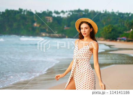Attractive young woman in a polka dot dress and straw hat strolls on a sandy beach by a calm sea with tropical foliage and beach huts in the background during a relaxed holiday retreat. 114141186