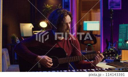 Young man songwriter playing guitar and creating music using equalizer program. Musician recording song in home studio, engineer mixing and mastering pop rock tracks. Camera A. Young man songwriter playing guitar and creating music using equalizer program. Musician recording song in home studio, engineer mixing and mastering pop rock tracks. Camera A. 114143391