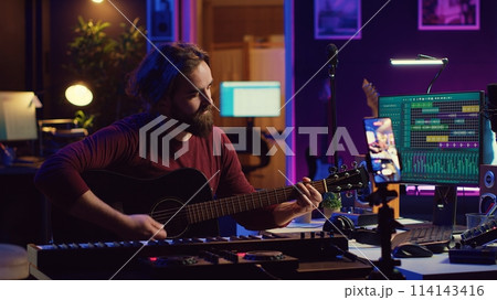 Skilled musician teaching how to play acoustic guitar online, creating internet tutorial to give assistance with musical instrument. Artist songwriter explaining accords and notes strings. Camera A. Skilled musician teaching how to play acoustic guitar online, creating internet tutorial to give assistance with musical instrument. Artist songwriter explaining accords and notes strings. Camera A. 114143416