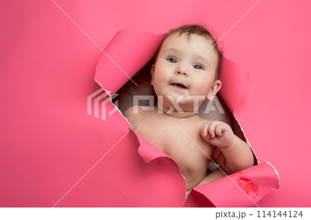 Cute Caucasian newborn baby boy peeks out of a hole in a paper pink background. 114144124