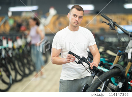 Young man choosing bicycle and checking bike handlebar quality in sport store Young man choosing bicycle and checking bike handlebar quality in sport store 114147395