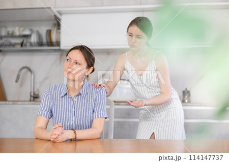Offended mother sitting at the kitchen table, daughter standing behind 114147737