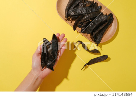 A beauty hand lifting some black locust pods on palm hand, a rectangle dish contains black locust fruit placed on yellow table. Photo was taken from top view, space for text 114148684