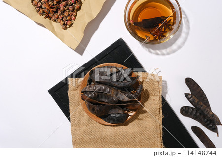 A dish of black locust fruits placed on sackcloth towel is spread on a rectangle black tray next to a glass bowl with brown liquid and some dried rose buds. Space for advertising or designing 114148744