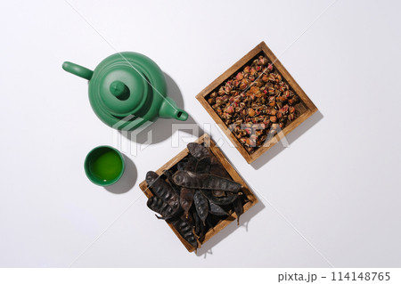 Top view photo with a green tea pot, a cup of tea, wooden tray of dried rose buds and a tray of black locust fruits placed side by side on white background. Space for text or designing 114148765