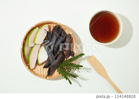 White background against a rattan tray with black locust seed pods and grapefruit peel on the left side and a bowl of brown liquid on the right. Copy space with view from above White background against a rattan tray with black locust seed pods and grapefruit peel on the left side and a bowl of brown liquid on the right. Copy space with view from above 114148928