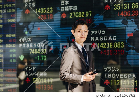 世界の株価が映る電光掲示板の前で笑顔で立つカメラ目線の30代の男性 114150862