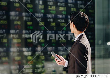 電光掲示板に映るリアルタイム株価ボードを見てスマホで取引をする30代のスーツ姿の日本人男性の後姿 電光掲示板に映るリアルタイム株価ボードを見てスマホで取引をする30代のスーツ姿の日本人男性の後姿 114150864