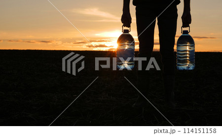 A man holds two bottles of drinking water, stands in a field at sunset A man holds two bottles of drinking water, stands in a field at sunset 114151158