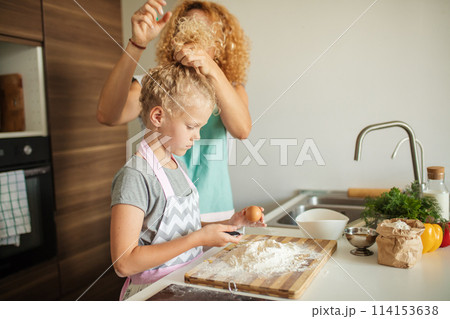 Little girl learn to make dough and her caring mother tying her hair up. 114153638