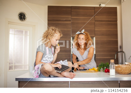 Little girl and her daughter baking cookies at home, reading recipe on laptop. Little girl and her daughter baking cookies at home, reading recipe on laptop. 114154256