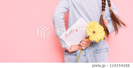 Little girl holding drawn card and flower behind her back for Father's Day holiday in front of pink background 114154288