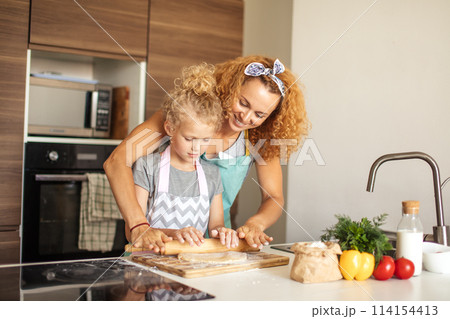 Beautiful young mom and her cute little daughter rolling out dough at home. 114154413