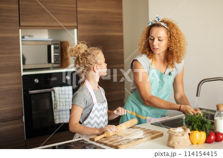 Beautiful young mom and her cute little daughter rolling out dough at home. Beautiful young mom and her cute little daughter rolling out dough at home. 114154415