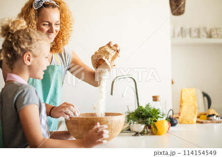 Little girl and her mother sprinkling flour into a bowl and smiling while baking 114154549