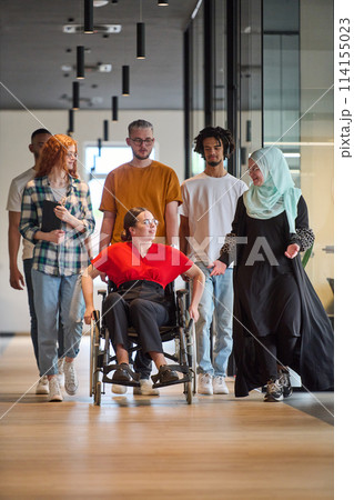 A diverse group of young business people walking a corridor in the glass-enclosed office of a modern startup, including a person in a wheelchair and a woman wearing a hijab 114155023