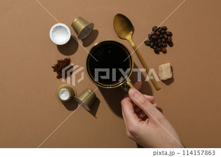 Coffee capsules, coffee beans, sugar, spoon and cup in hand on brown background, top view 114157863