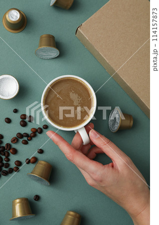 Coffee capsules and beans, cup with coffee drink in hand and box on green background, top view Coffee capsules and beans, cup with coffee drink in hand and box on green background, top view 114157873