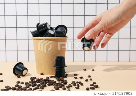 Black coffee capsules in paper cup, hand and coffee beans on light background Black coffee capsules in paper cup, hand and coffee beans on light background 114157902