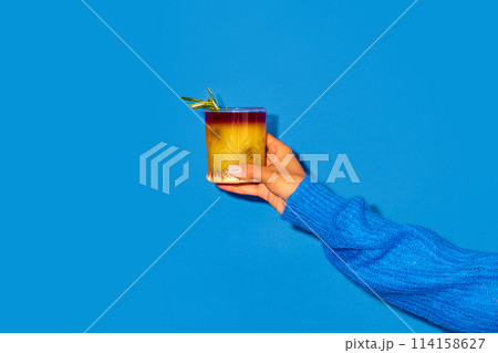Female hand holding cocktail with orange juice against blue background. New York sour traditional cocktail Female hand holding cocktail with orange juice against blue background. New York sour traditional cocktail 114158627