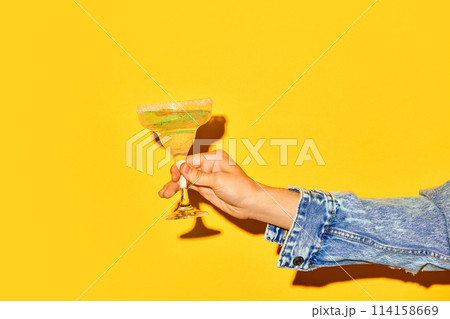 Male hand holding sweet and sour margarita cocktail with slice of orange against bright yellow background Male hand holding sweet and sour margarita cocktail with slice of orange against bright yellow background 114158669