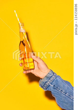 Human hand holding bottle with lemonade with straw against bright yellow background 114158696