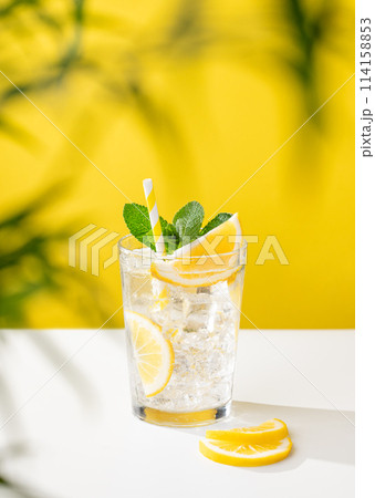 A glass of cold lemonade with lemon, mint and ice on a yellow and white background with a palm leaf. A glass of cold lemonade with lemon, mint and ice on a yellow and white background with a palm leaf. 114158853