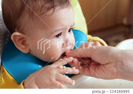 Portrait of little baby boy eating food. Baby with a spoon in feeding chair. Cute baby eating first meal 114160995