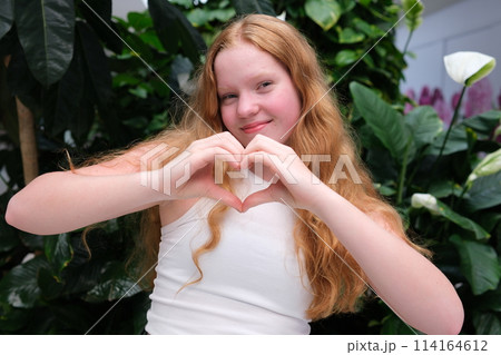 Beautiful red-haired, young woman, girl smiling, making a heart gesture against a background of greenery. High quality photo Beautiful red-haired, young woman, girl smiling, making a heart gesture against a background of greenery. High quality photo 114164612